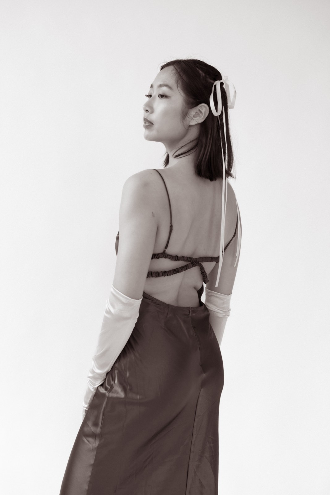 Sepia-toned photo of Nikki from behind in a satin skirt and long opera gloves, a ribbon tied in her hair, looking over her shoulder