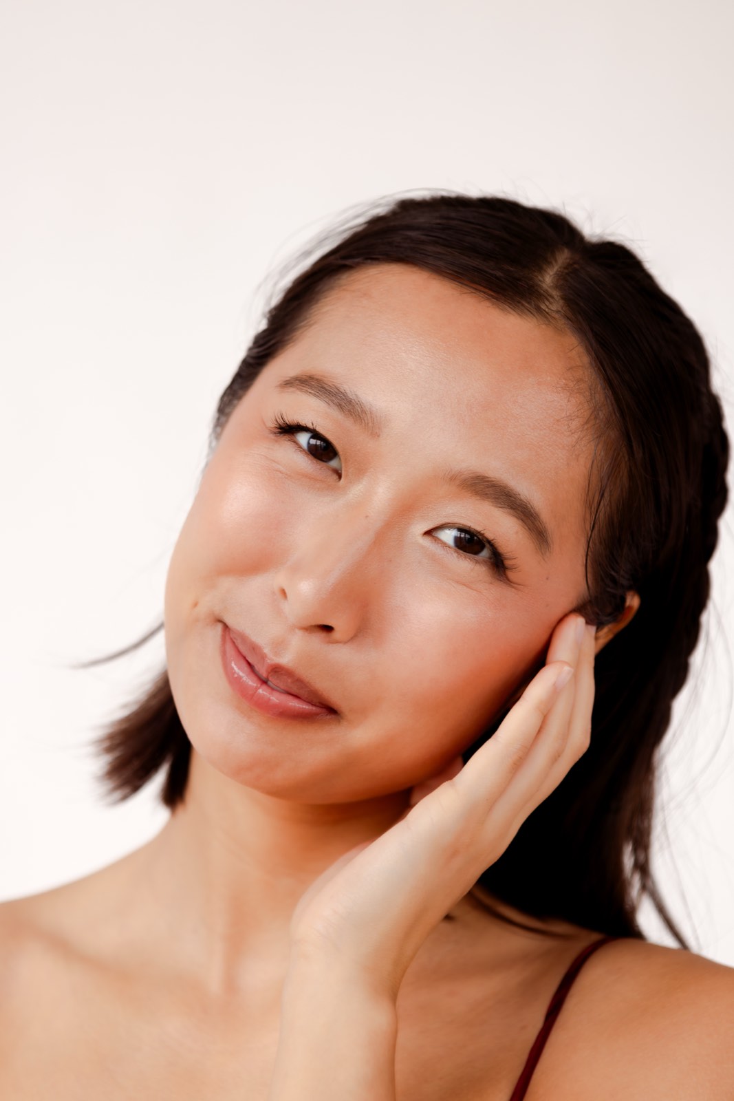 Nikki resting her cheek on one hand, smiling softly at the camera in a red strap top against a cream backdrop