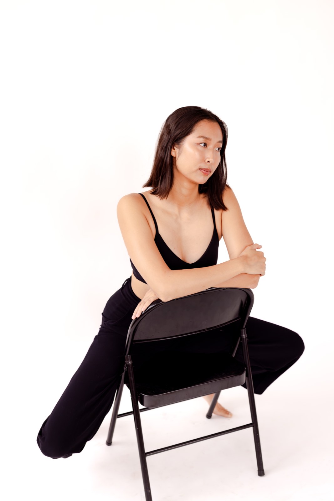 Nikki seated backwards on a black folding chair, arms crossed over the backrest, in a black cami and loose trousers