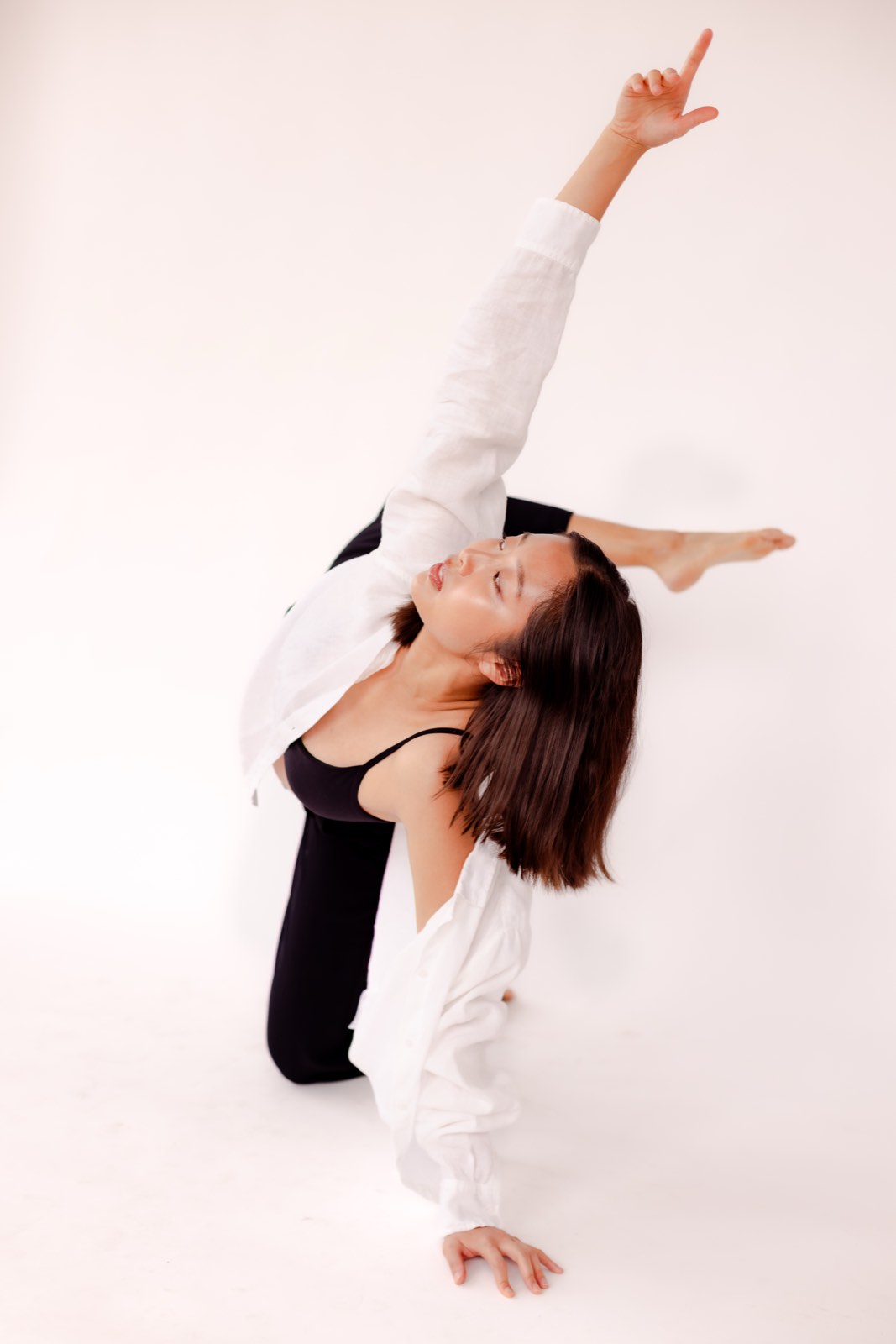 Nikki kneeling in a deep backbend with one arm reaching upward, in an open white shirt over black, on a white studio backdrop