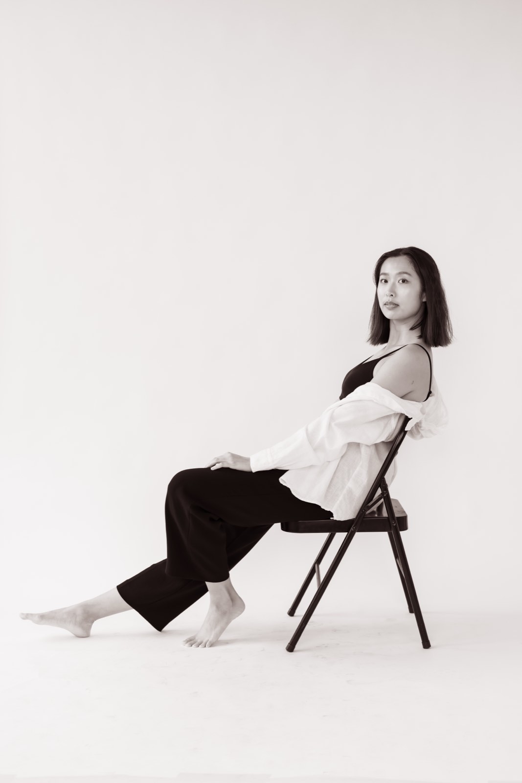 Sepia photo of Nikki reclining on a folding chair in an open white shirt over black, legs stretched out
