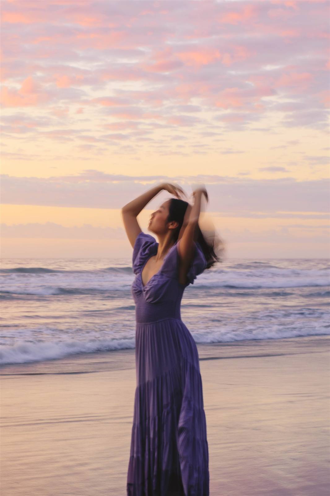 Nikki on the shoreline at sunset with both arms raised to her hair, in a long purple dress against a pink sky