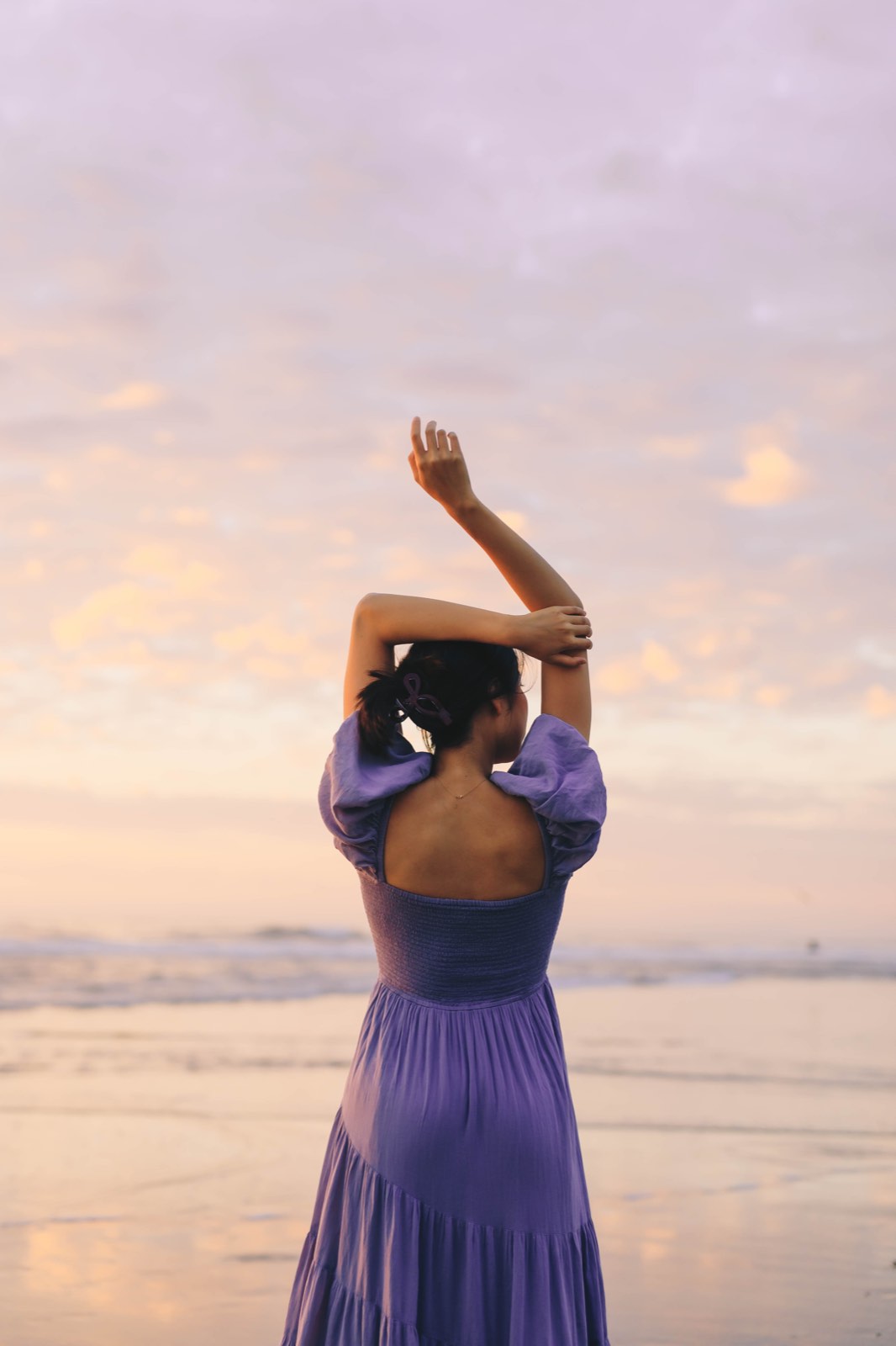 Nikki on the beach at sunset, seen from behind with both arms raised, wearing a lavender tiered dress under a pink and gold sky