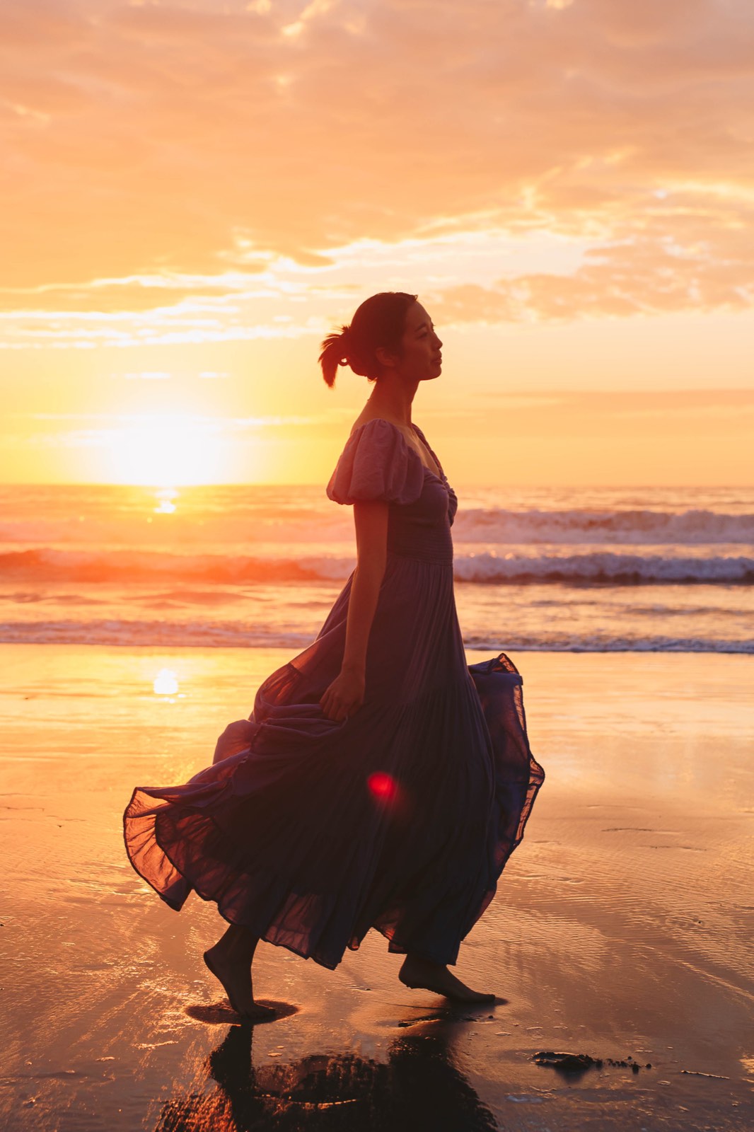Nikki silhouetted against a blazing orange sunset, walking barefoot across wet sand and holding out the skirt of a purple dress