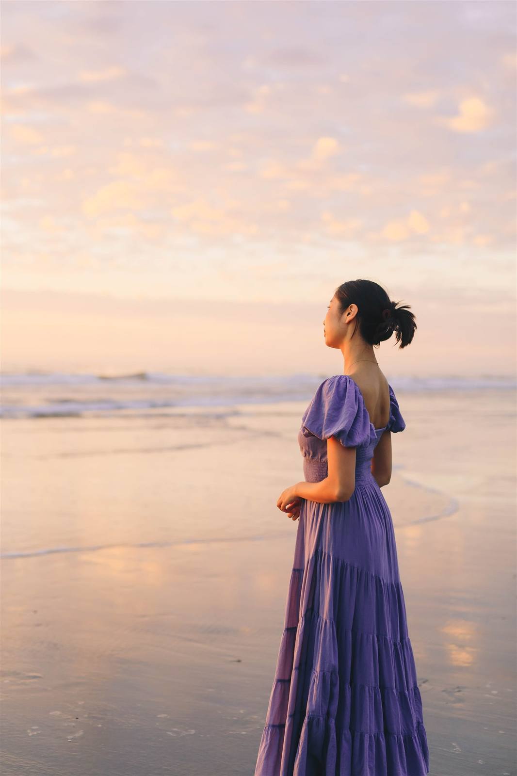 Nikki standing in profile at the water's edge in a purple tiered dress, looking out over a pale golden sea