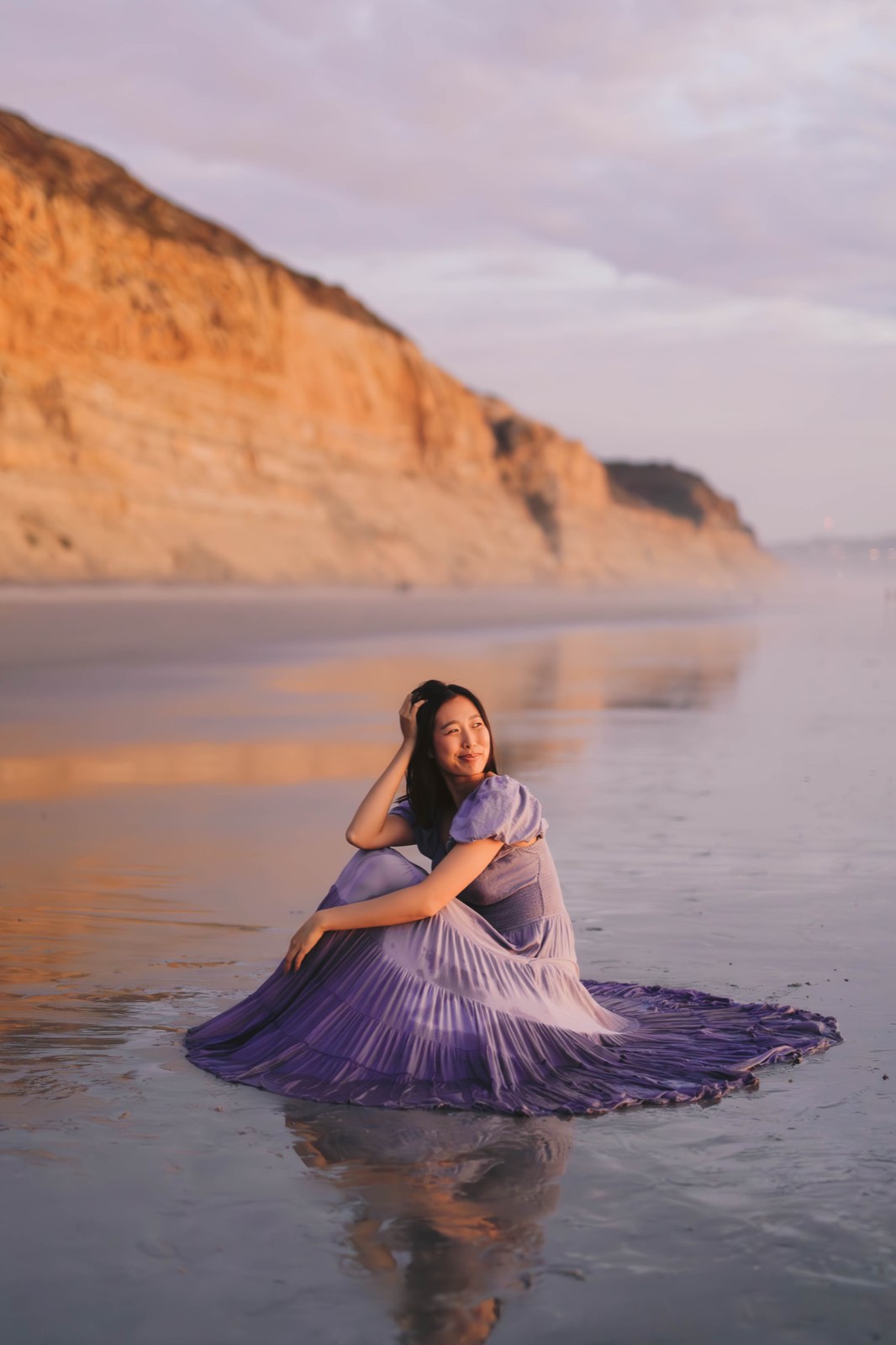 Nikki seated on wet reflective sand with her purple pleated dress fanned around her, sandstone cliffs rising behind in soft evening light
