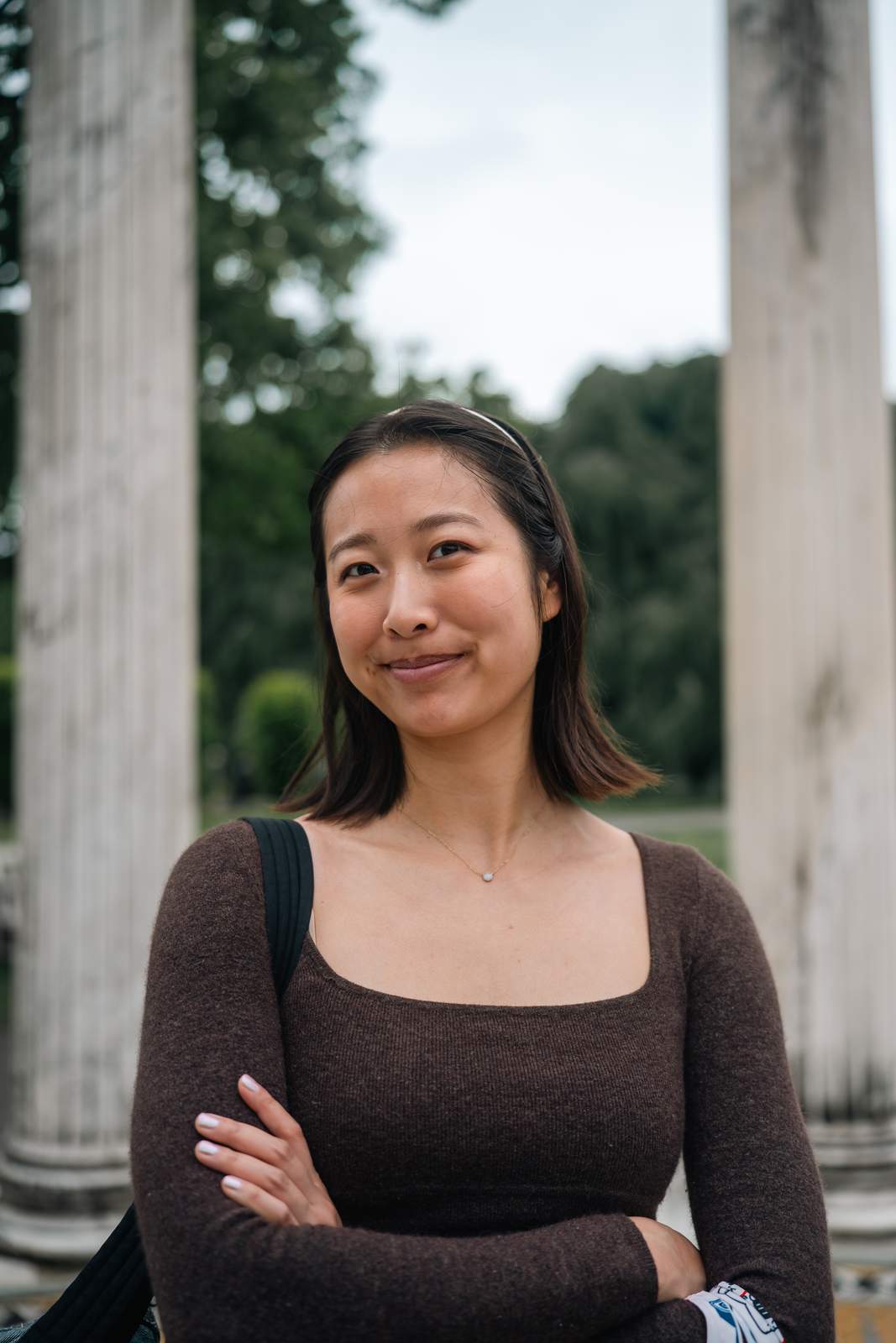 Nikki smiling between two weathered stone columns in a garden, arms folded, in a brown knit top