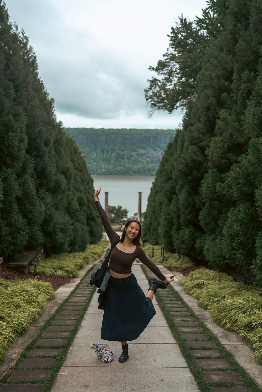 Nikki balancing on one leg with an arm raised, on a path between tall hedges looking out to the Hudson River