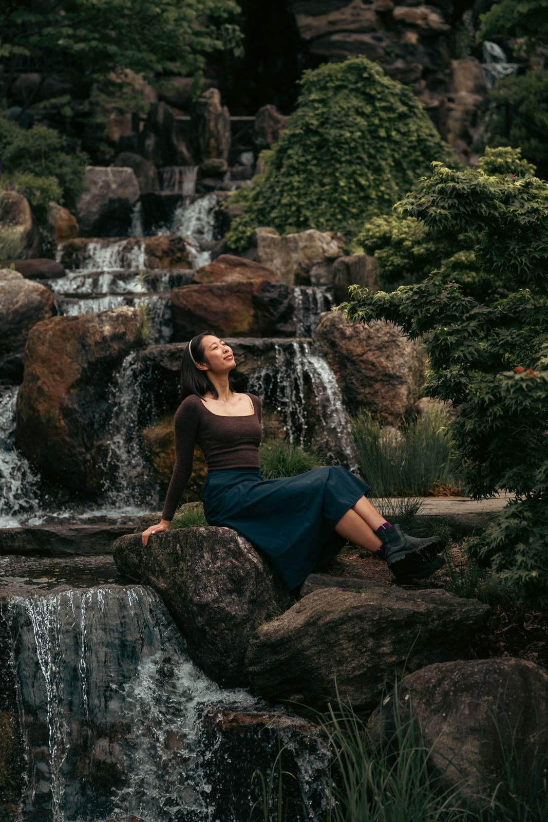 Nikki seated on a rock beside a cascading garden waterfall, face tilted up, in a brown top and navy skirt