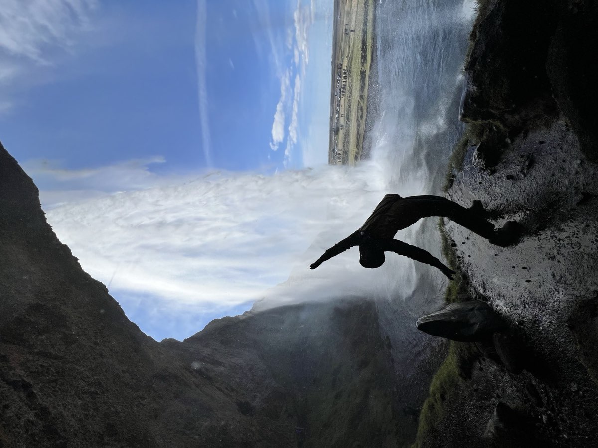 Nikki silhouetted behind a curtain of falling water at an Icelandic waterfall, blue sky beyond