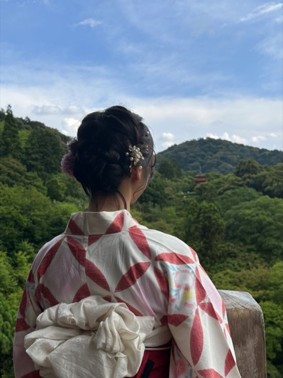 Nikki from behind in a red and white yukata, looking out over forested hills toward a distant pagoda