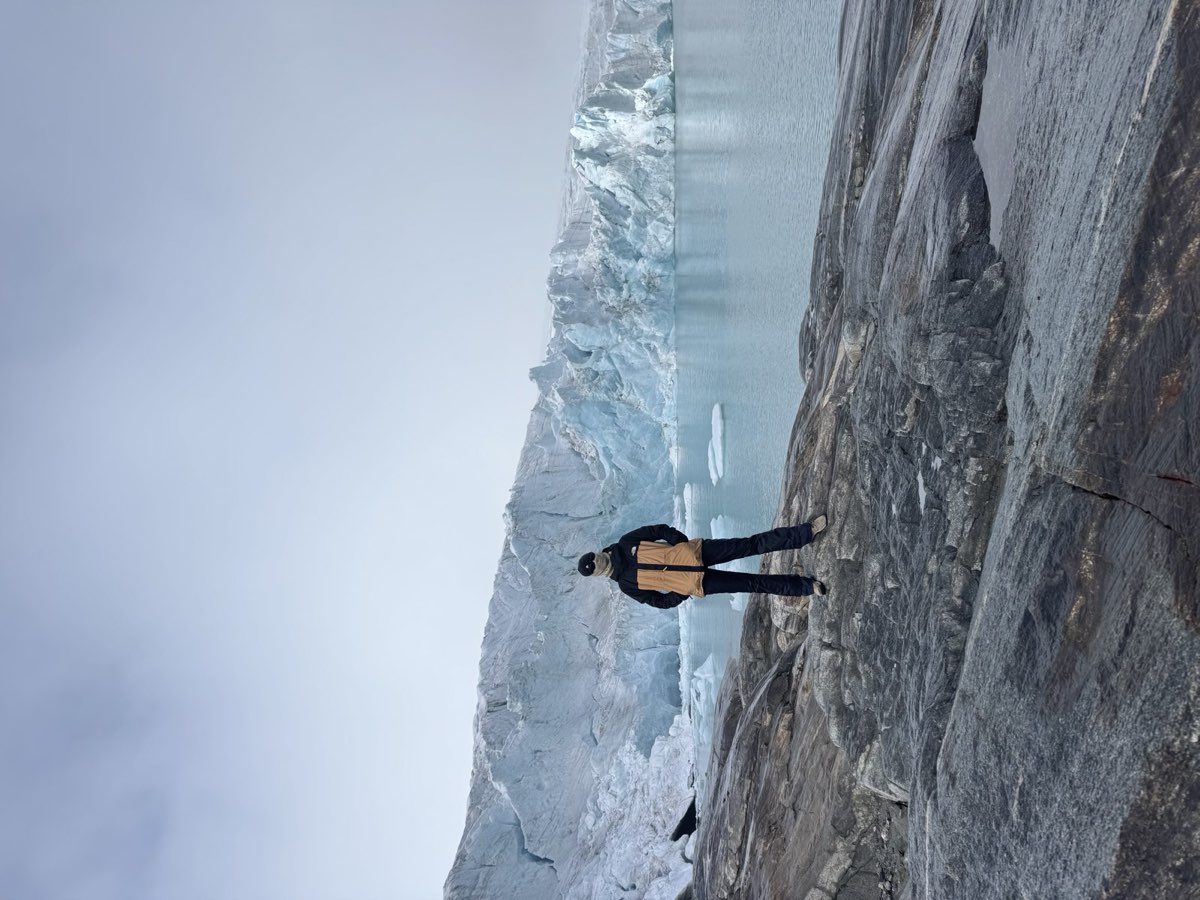 Nikki standing at the edge of a rock ledge overlooking a pale blue glacier and its meltwater lake