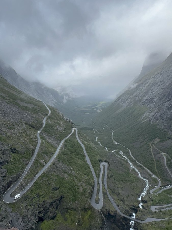 A mountain road switchbacking down a steep green Norwegian valley beside a waterfall, under low cloud
