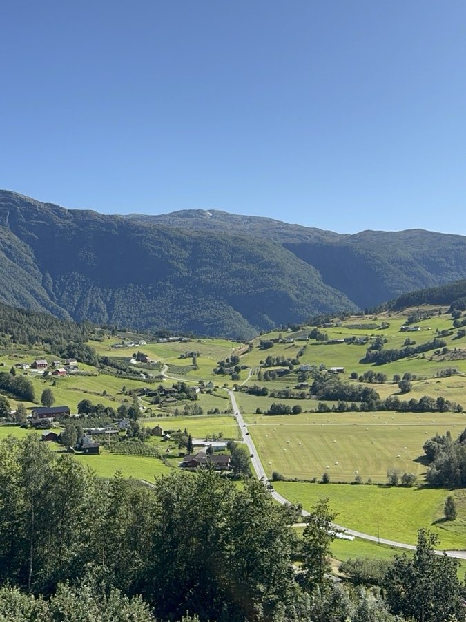 A sunlit Norwegian valley of green farmland, red barns and hay bales below forested mountains