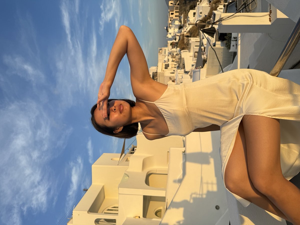 Nikki in a cream slip dress on a whitewashed terrace, hand raised to her face, blue sky behind