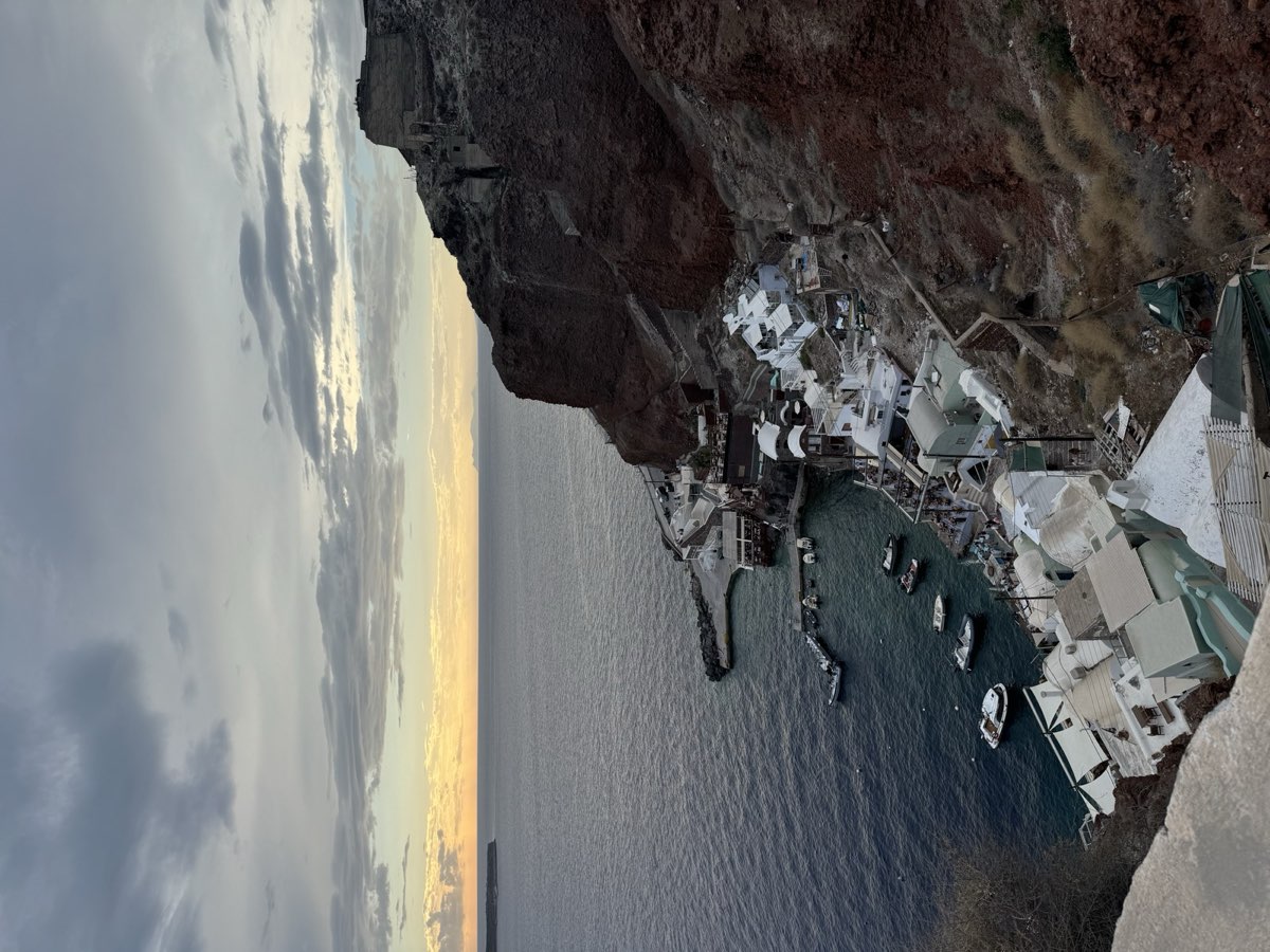 Whitewashed houses tumbling down a cliff to a small harbour of moored boats at dusk, Santorini