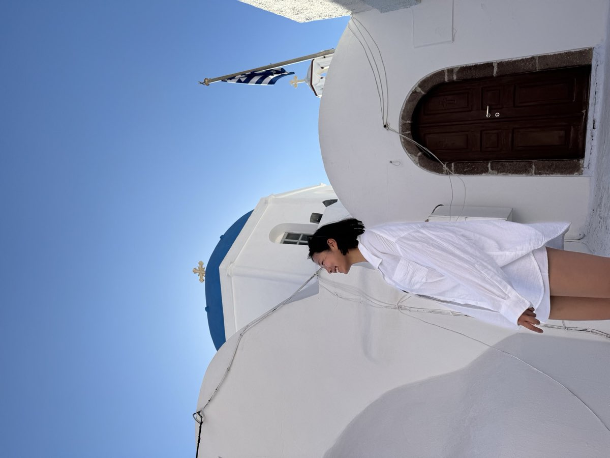 Nikki in a white shirt beside a blue-domed whitewashed church, a Greek flag against deep blue sky