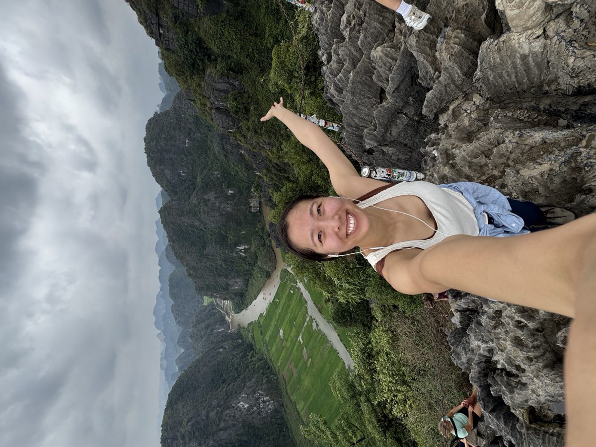 Nikki taking a selfie at a summit with one arm raised, above limestone karst peaks and a winding river
